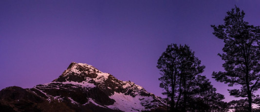 Will was waiting for us, explaining that after having taken a jaunt up to Travers Saddle with Nina and Ian, they had decided to head down valley and would rejoin us in two days. While it would have been nice to have their company, that evening we savoured the luxury of having the entire hut to ourselves, despite it being Labour weekend.  
Next morning dawned crisp and clear – better than the morning of our climb, despite the weather forecast suggesting otherwise. Simon, Caro and I headed for Travers Saddle and soon discovered that there had also been a freeze overnight. With the clarity of hindsight we realised that this should have been our summit day. “Buggar! Ah well” we thought, “might as well make the most of the situation and go check out the grade three route up from the Saddle, along the South Ridge“. This image - The eastern aspect of Mt Travers (2,338m) at dawn, viewed from behind Upper Travers Hut.