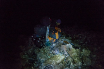 All of us were fully clothed in almost everything we had, with boots still on. I was alone in a bivvy bag trying to control violent bouts of shivering, pondering how we could last like this for 13 hours. Simon and Caro shared a bothy bag slightly uphill behind a protruding boulder. As the minutes dragged by, frequent bursts of laughter drifted my way between wind gusts and the ominous rattling and thudding of falling rocks. I couldn’t see even the slightest shred of humour in our situation, yet they seemed to be having a rip-roaring time. Actually, they weren’t. Caro confessed later ‘It was a case of laugh or cry.’
At about 11.30pm the wind eased, clouds cleared and a still near-full-moon lit things up. Now we could see that Tasman Glacier was not far down at all. ‘Let’s get out of here while we still can’ exclaimed Simon. No argument there! I set off first to get well ahead while they stashed their gear, so as to avoid rock fall from them as they descended.  
This image - About to depart our 1,050m bivvy site at 11.30pm.